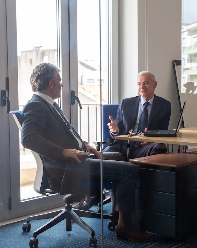 Friendly mature business partners meeting in office, sitting at workplace with laptop and talking. View through glass wall. Business portrait concept